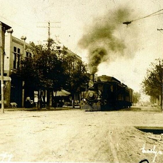 Train on Eveline, 1906, George Barrowclough Black and white image of a steam train going down a street with buildings on either side.