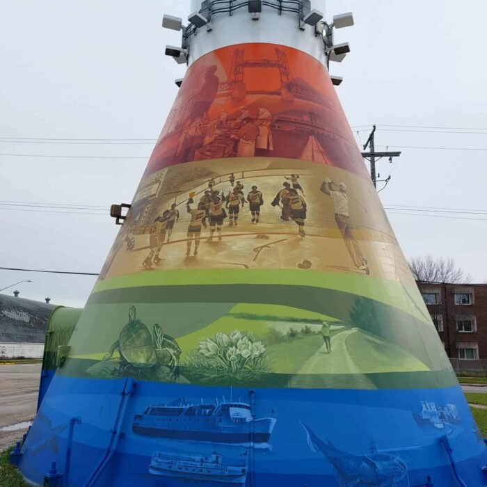 Colour image of a mural painted on the base of the Selkirk Water Tower. There are four colours used, red, orange green, and blue. Each band of colour has various scenes of people, places, and animals