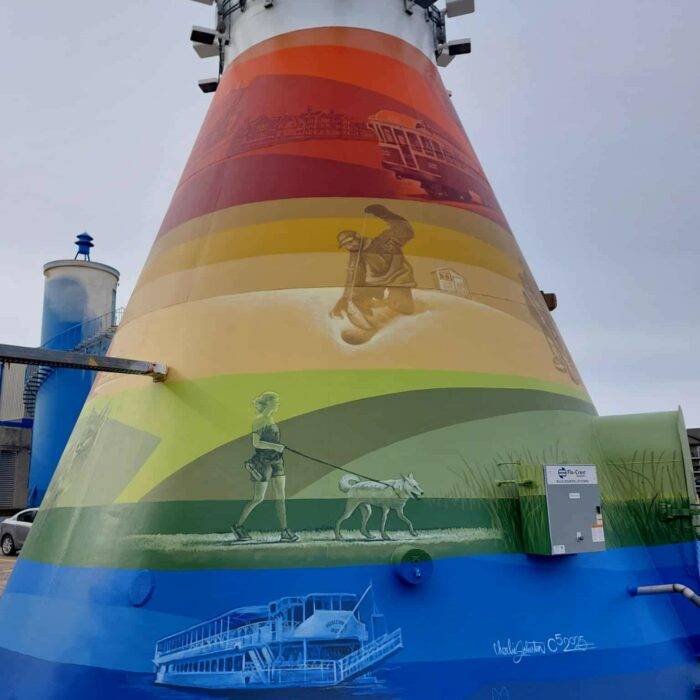 Colour image of a mural painted on the base of the Selkirk Water Tower. There are four colours used, red, orange green, and blue. Each band of colour has various scenes of people, places, and animals