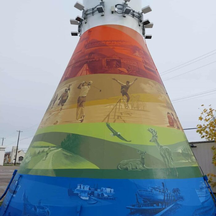 Colour image of a mural painted on the base of the Selkirk Water Tower. There are four colours used, red, orange green, and blue. Each band of colour has various scenes of people, places, and animals