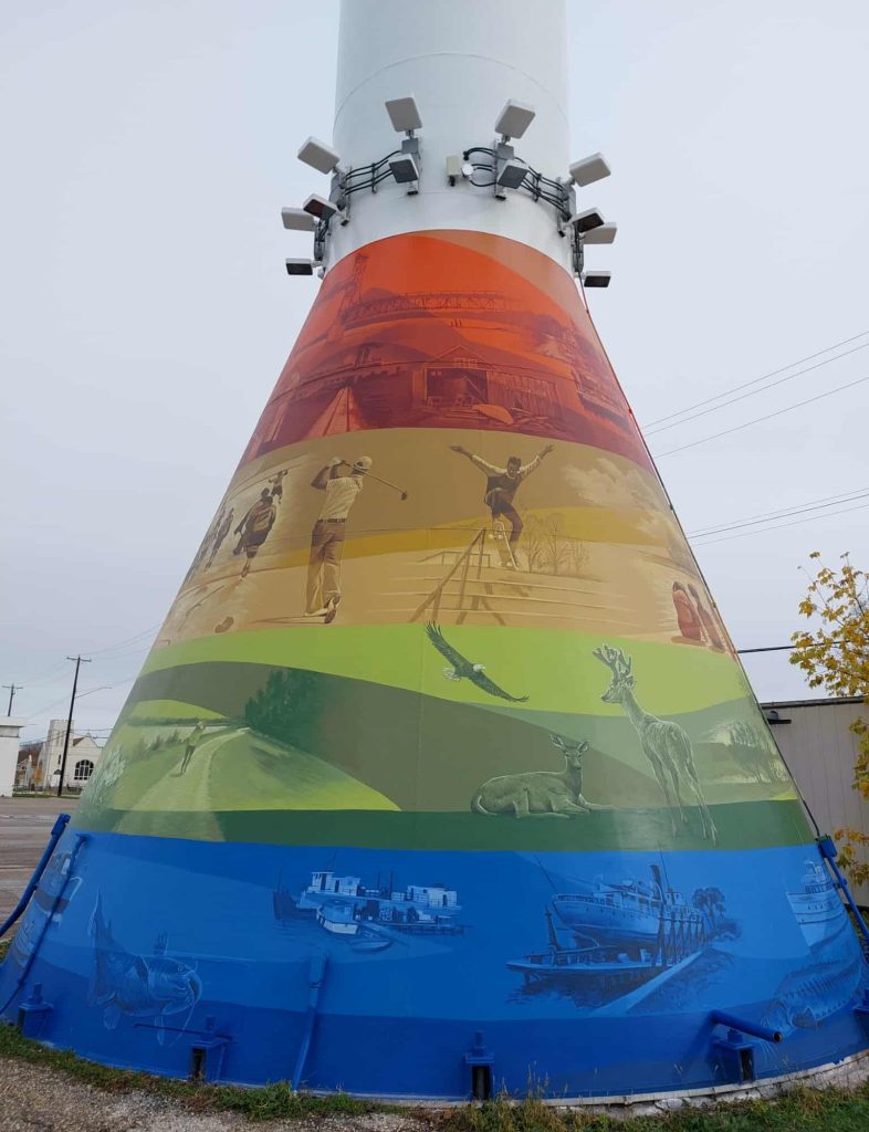 Colour image of a mural painted on the base of the Selkirk Water Tower. There are four colours used, red, orange green, and blue. Each band of colour has various scenes of people, places, and animals