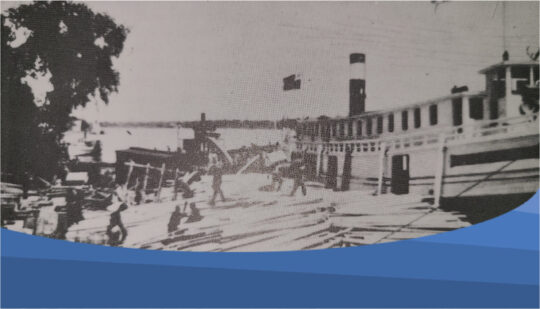 Photo of lumber transport in Selkirk The photo shows a historic shot of lumber being loaded from the red river onto the shore of Selkirk.