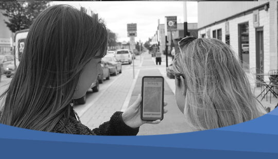 Selkirk Museum BW Photo taken from behind two people looking at the Selkirk Museum website on a phone. They are standing on the sidewalk of Manitoba Avenue.