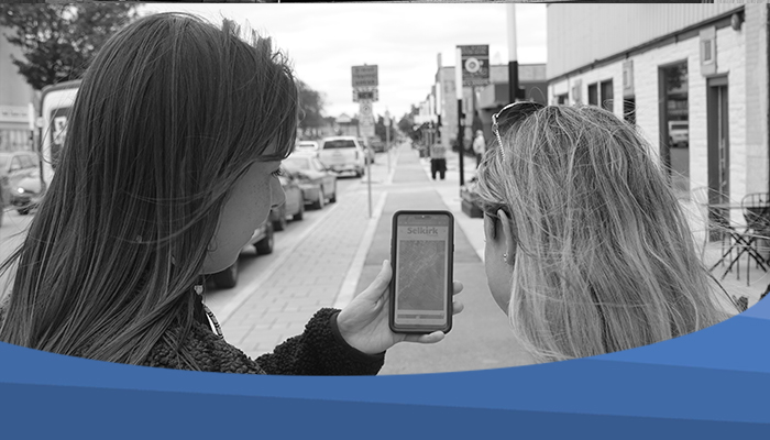 Selkirk Museum BW Photo taken from behind two people looking at the Selkirk Museum website on a phone. They are standing on the sidewalk of Manitoba Avenue.