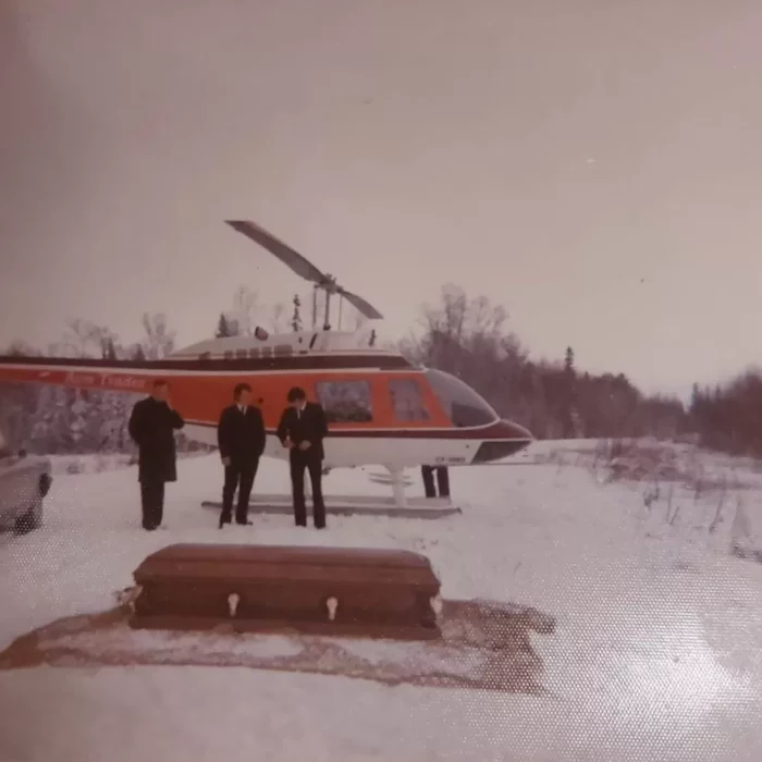 Heli Lifting a Casket at Loon Straights, Date Unknown, Wes Gilbart Helicopter on a snow with three people standing in front of it. A casket is in front of them.