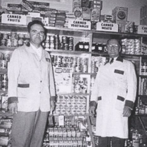Black and white portrait of two men standing in front of canned goods