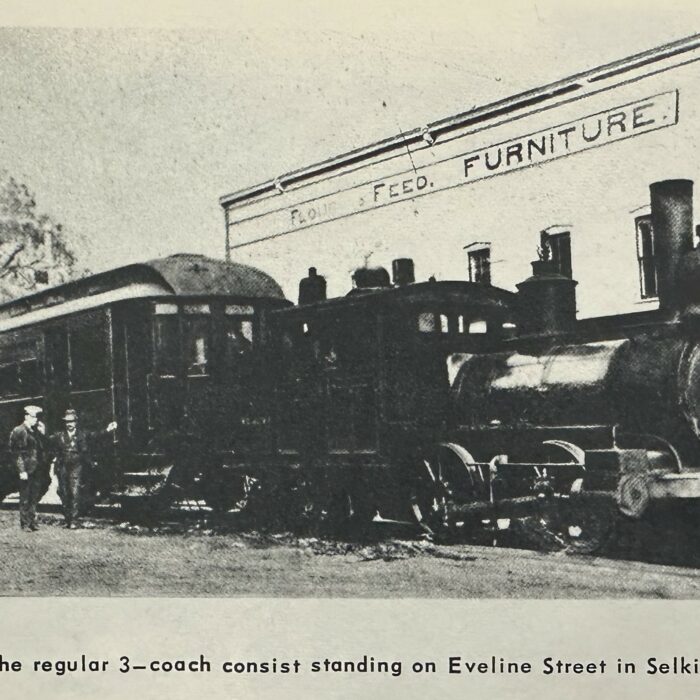 Steam engine at a standstill on Eveline St. Selkirk MB. there is a building in the background and a person standing beside the train