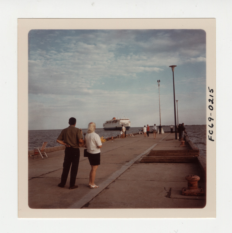 Colour image of people standing on a dock. There is a large boat in the distance.
