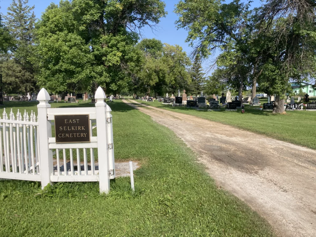Photo of white fence around a cemetery with headstones and trees.