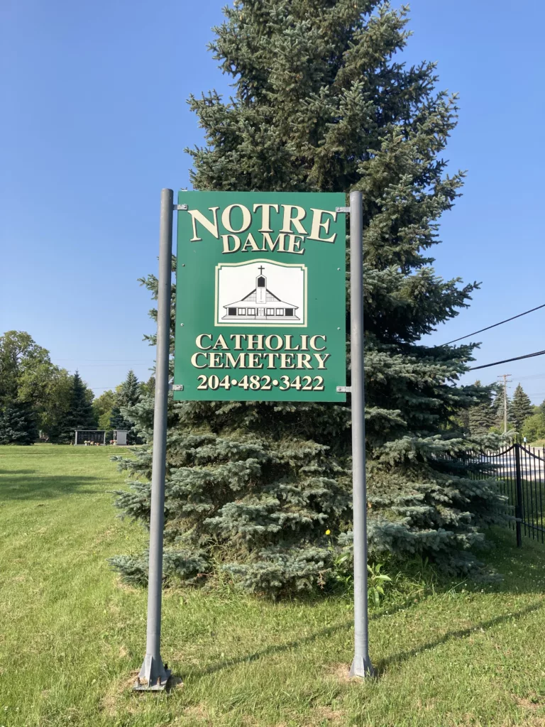 Photo of green sign for the cemetery. Behind the sign is a large evergreen tree.