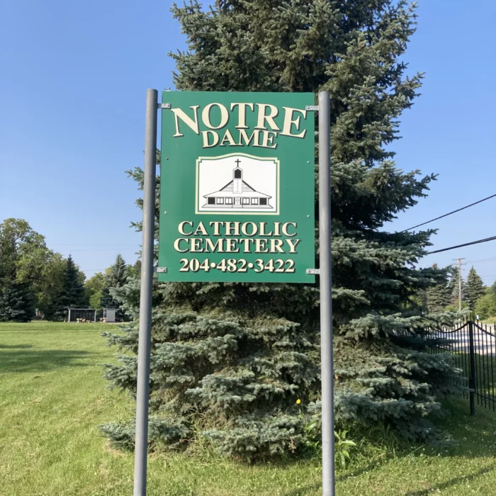 Photo of green sign for the cemetery. Behind the sign is a large evergreen tree.