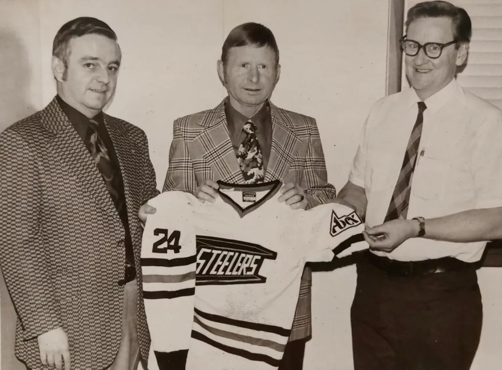 Three men wearing suits and holding a hockey jersey with the word Steelers on it, in this black and white photo.