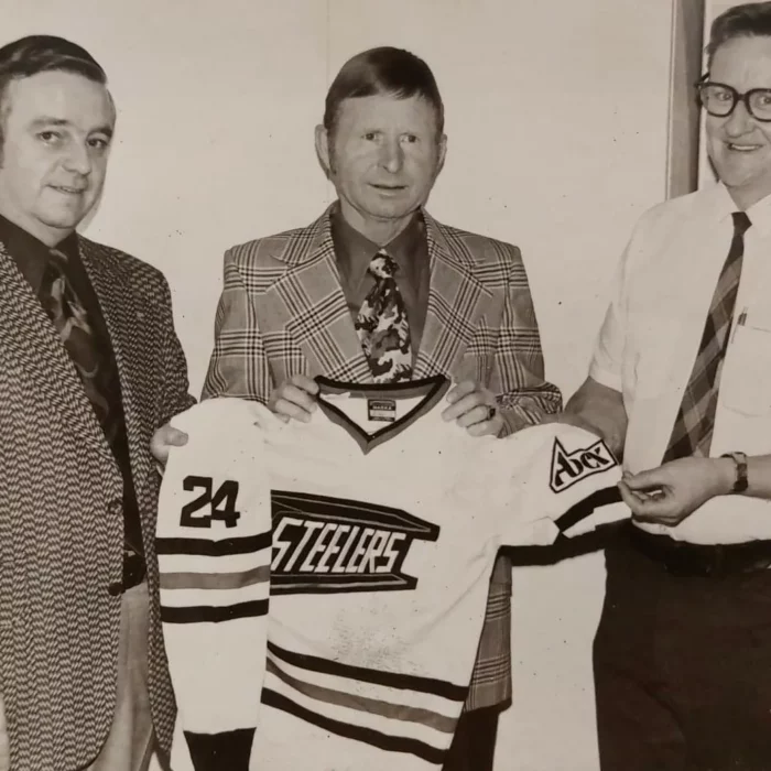 Three men wearing suits and holding a hockey jersey with the word Steelers on it, in this black and white photo.