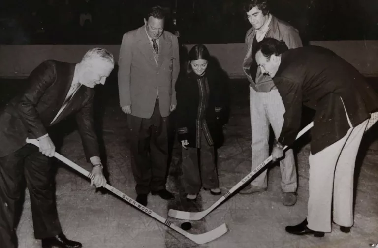 Black and white photo of a woman dropping a hockey puck and two men facing off with hockey sticks.