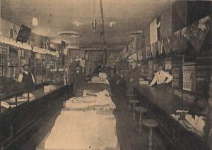 Inside the store of Shepard Peers and Co. Three men at looking at the camera with stacked shelves behind them