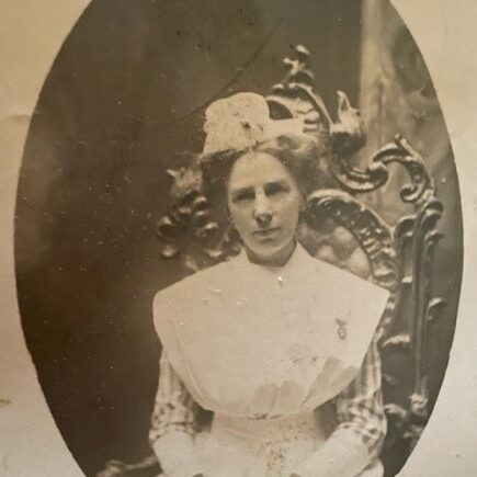 Miss Ada Janet Ross, c1900s, Joen Hadfield Black and white photo of a young woman seated on an ornate wood carved chair. She is wearing a nurses uniform, a broach, and a hat. She looks just left of the camera and has her hands clasped on her lap