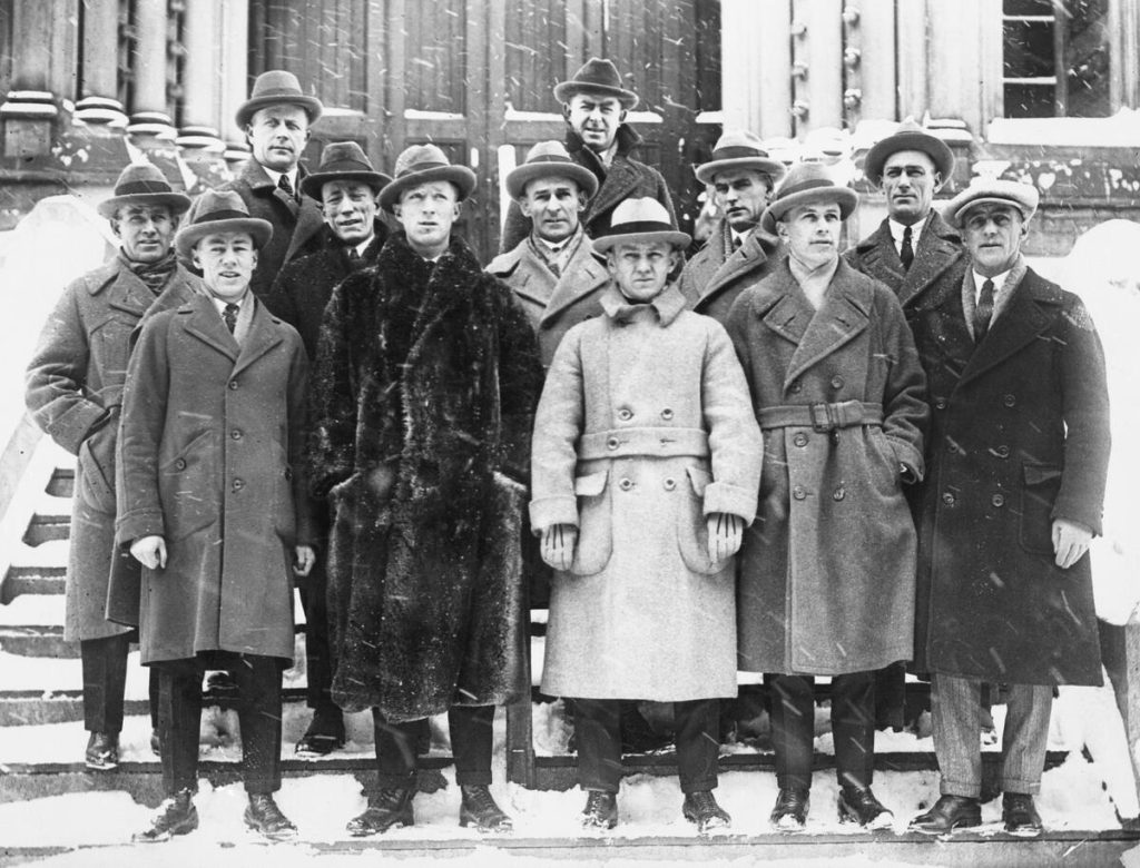 Black and white image of a group of men posing for a photo outside the front door of a building during a snowfall