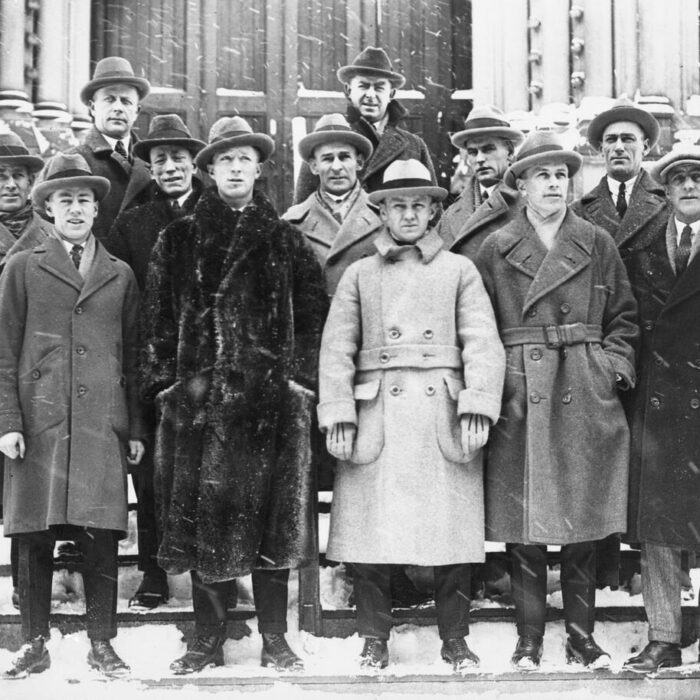 Black and white image of a group of men posing for a photo outside the front door of a building during a snowfall
