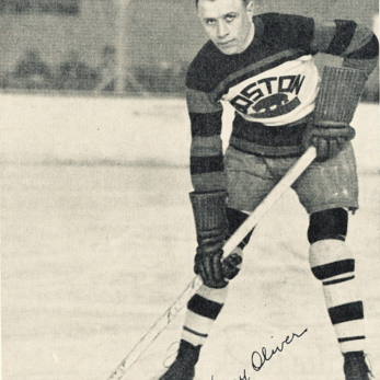 Black and white image of Harry Oliver posing on the hockey ice surface wearing his Boston Bruins jersey and holding a hockey stick
