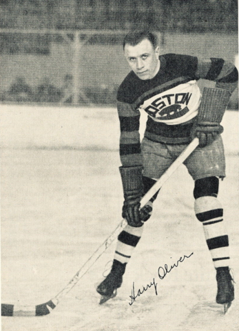 Black and white image of Harry Oliver posing on the hockey ice surface wearing his Boston Bruins jersey and holding a hockey stick