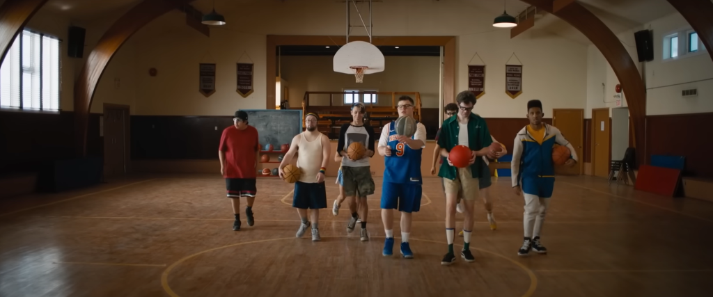 A group of basketball players walks towards the camera. A few hold basketballs. At the back of the hall is a basketball hoop
