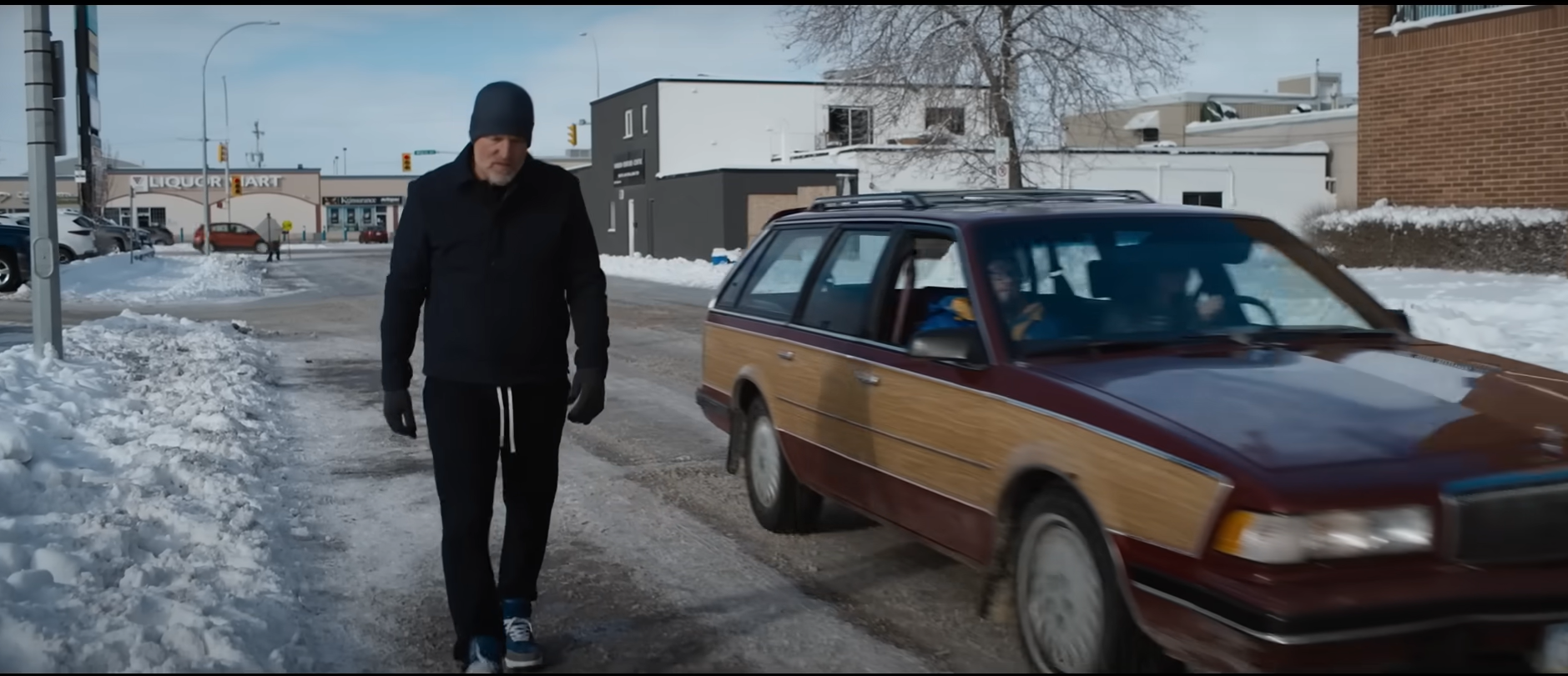 Image of a man walking down the road. A brown station wagon is beside him