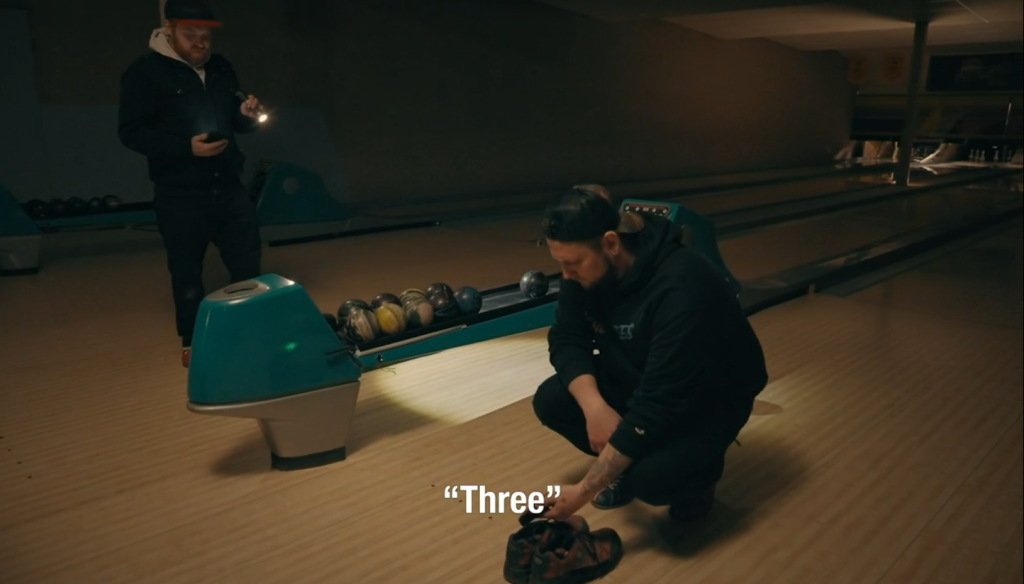 Image shows a man crouched beside a bowling ball cannon touching some shoes. A second man is standing on the opposite side of the bowling ball cannon. He is holding a flashlight and pointing it towards the shoes