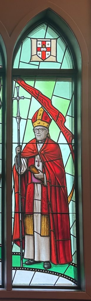 Edward Scott stands holding a staff, he is wearing a red robe and a mitre
