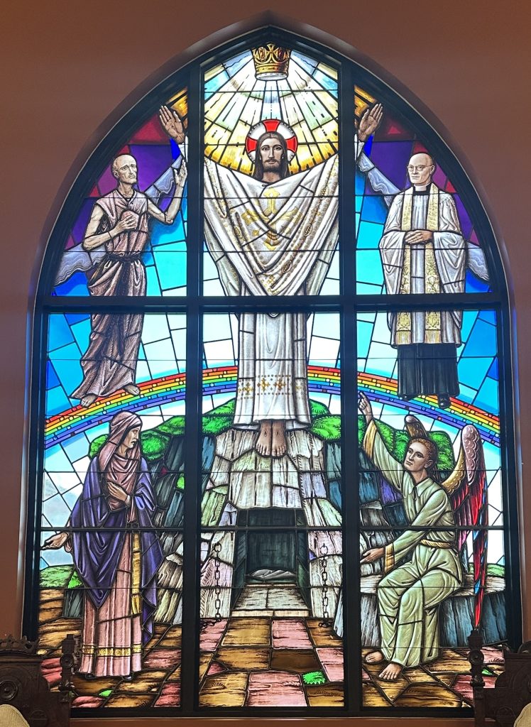 Large colourful window showing Christ in the center with four figures reaching up to him. The window is the brightest around Christ's head and directly behind him. There is a rainbow separating the top half of the window from the bottom half