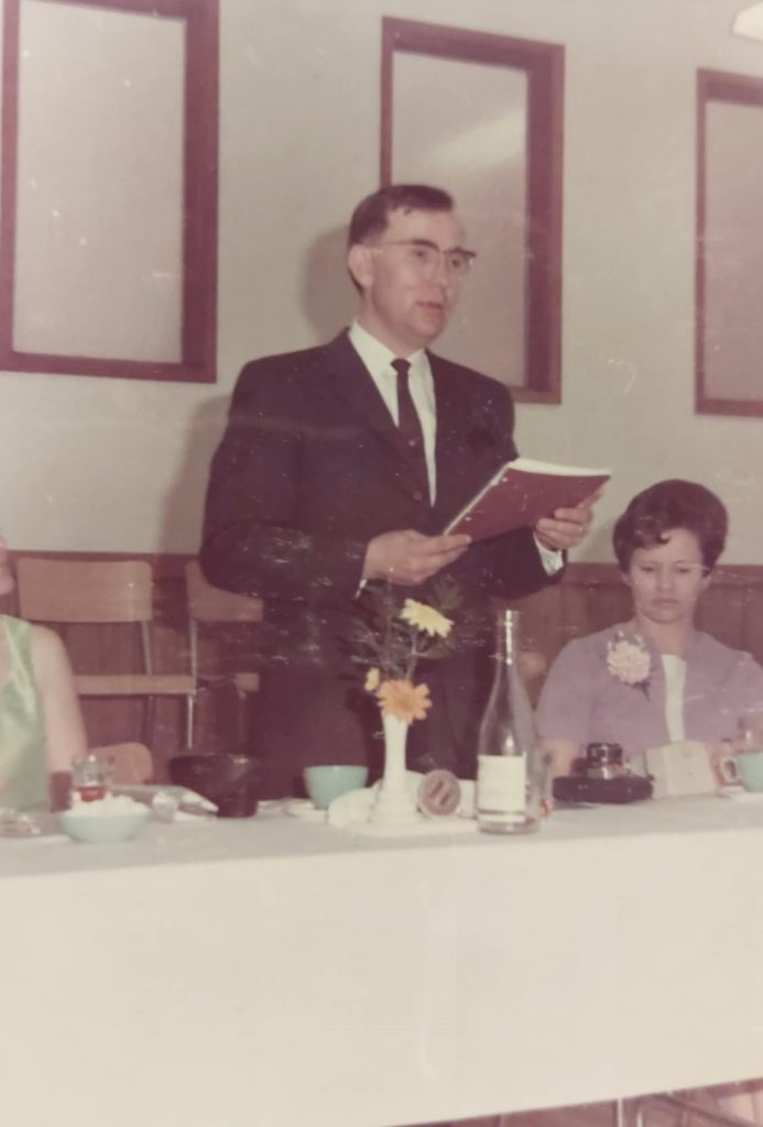 Image of Bill Rozecki standing at a table holding a book and speaking to the crowd