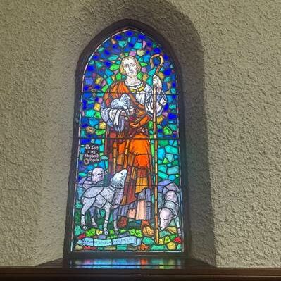 Stained glass window with Jesus wearing a red robe., holding a staff, and two sheep at his feet