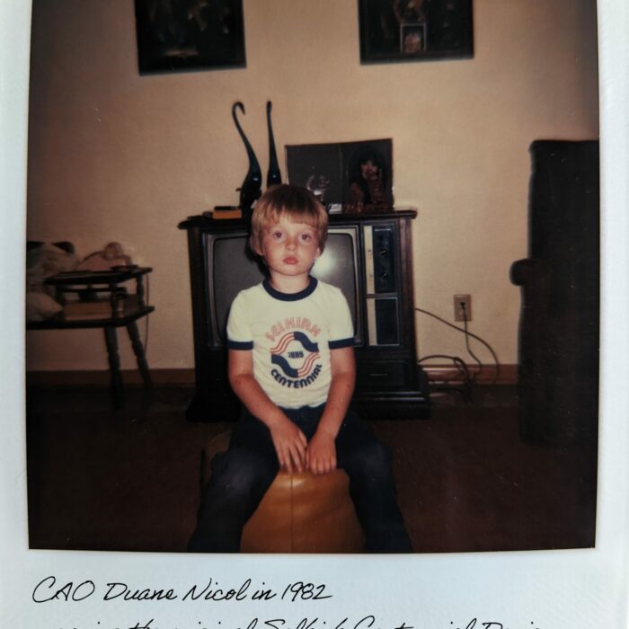 CAO Duane Nicol Wearing the Selkirk Centennial T-Shirt, 1982, Selkirk Museum Collection Colour polaroid image of a young boy seated a living room. He is wearing a white tee shirt which has the centennial logo on the front