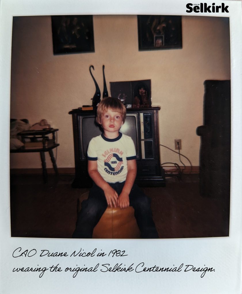 Colour polaroid image of a young boy seated a living room. He is wearing a white tee shirt which has the centennial logo on the front