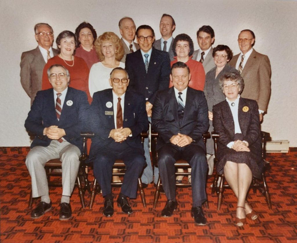 Colour photograph of a group of people seated and standing. They are in a room with white walls and red and black checkered floors