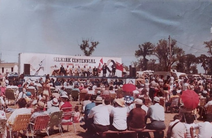 Colour image of the Centennial stage at Selkirk Park. A large group of people are clustered around the stage. The stage has a banner which reads 'Selkirk Centennial'