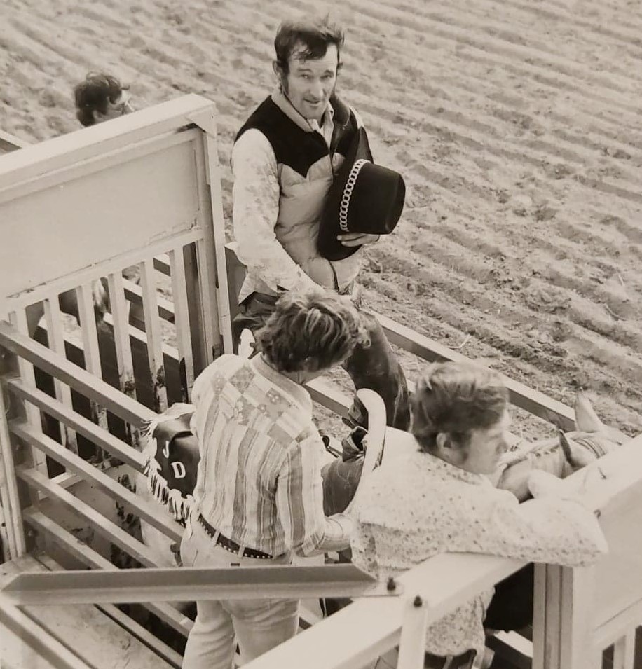 Black and white image of a man with a cowboy hat sitting on a cattle chute waiting to get on a horse