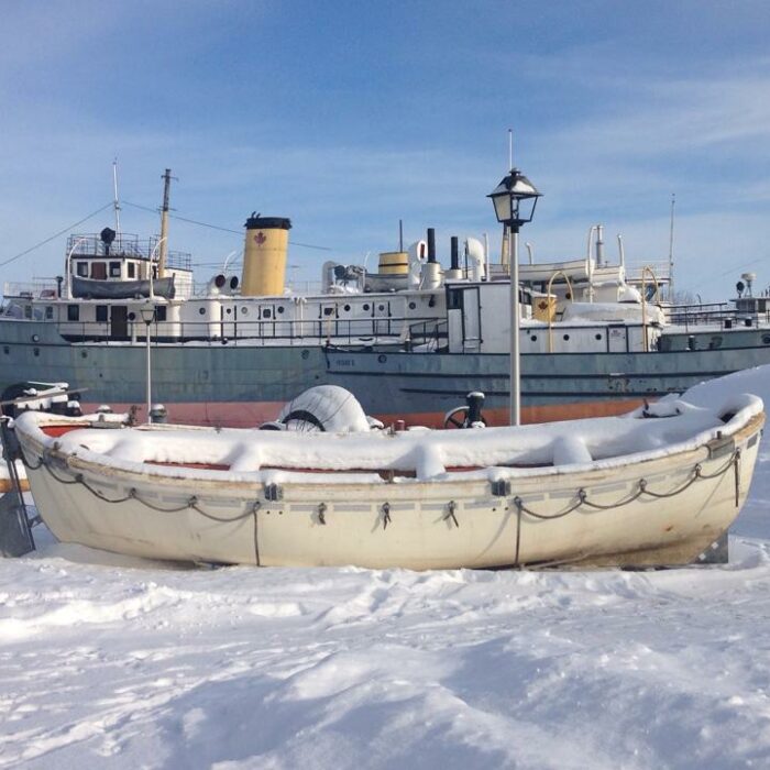 Lifeboat from S.S. Lord Selkirk, 2015, Marine Museum of Manitoba Colour photograph of the Bradbury, which is grey and orange, and a lifeboat from the Lord Selkirk. Both boats are docked on land and sit in the snow