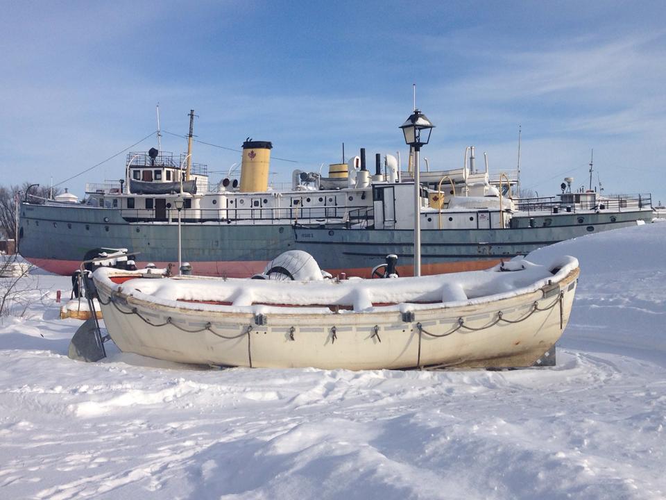 Colour photograph of the Bradbury, which is grey and orange, and a lifeboat from the Lord Selkirk. Both boats are docked on land and sit in the snow