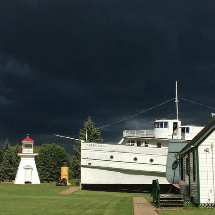 A colour image of a white wooden boat and a small white lighthouse in the background. Both are docked on the grass. The sky is a dark navy blue colour which looks like nighttime, but it is simply a huge thunderstorm