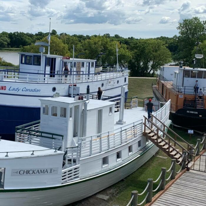 The Marine Museum, c2020, Marine Museum of Manitoba A colour image of two small white boats. Both are docked on the grass and one has a staircase leading to a wooden boardwalk