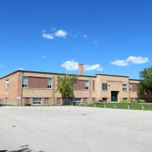 Colour photo of Daerwood school. The blue sky has a few small clouds at the center of the image. There are a few trees at the front of the school and a large paved driveway leading up to the entrance