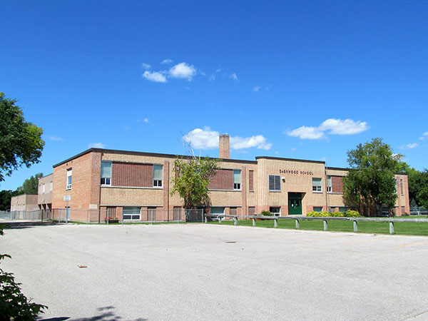 Colour photo of Daerwood school. The blue sky has a few small clouds at the center of the image. There are a few trees at the front of the school and a large paved driveway leading up to the entrance