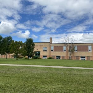 Colour image of Daerwood School. The blue sky is full of clouds. Trees are to the left of the photo frame and there is a large grassy space in front of the school. A sidewalk cuts across the image from left to right