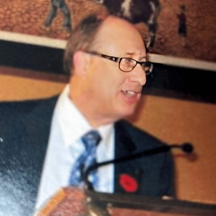 Founding Chairperson Lewis Wasel, 1995-2001, Selkirk District Community Foundation A man wearing narrow glasses and a black suit stands at a microphone stand