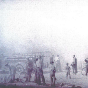 A black and white photo of firemen and a firetruck partly obscured by smoke