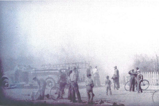 A black and white photo of firemen and a firetruck partly obscured by smoke