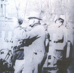 A black and white photo of three men standing. One has a bike. Stanley Dewar, the man in the center of the frame has his hand raised as he looks forward