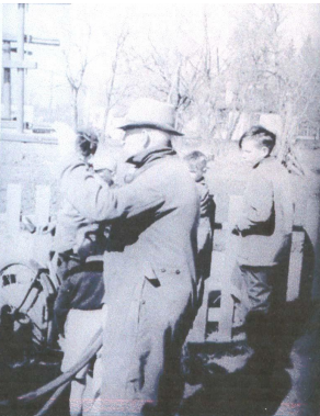 A black and white photo of three men standing. One has a bike. Stanley Dewar, the man in the center of the frame has his hand raised as he looks forward