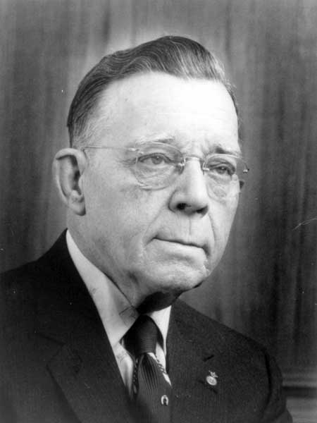 Black and white image of a man looking to the right. He is elderly and wears think framed glasses. He is wearing a black suit jacket, white shirt, and a black tie. He has short hair