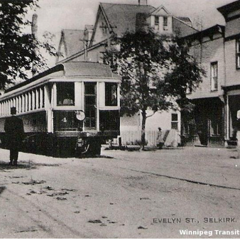 Black and white image of a street car driving down Eveline Street. A man stands to the left of the car